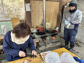 一般社団法人　甲賀ナイスローカルカンパニー　活動のようす画像　　　小屋の中に据えられたかまどで薪が燃えて大きな鍋を煮ている。手前では女性が肉を切っている